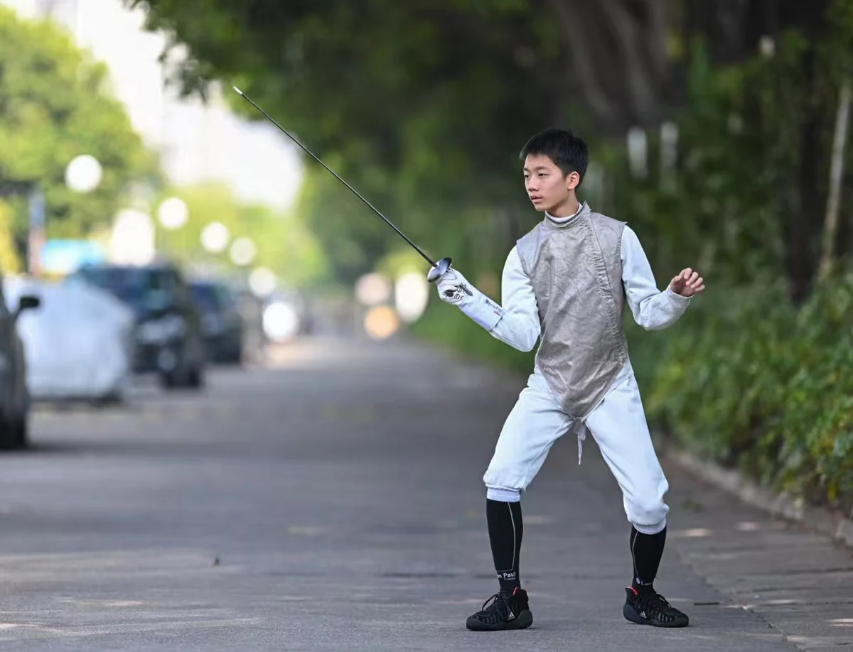 Steven in a fencing uniform holds a foil while standing on a tree-lined path.