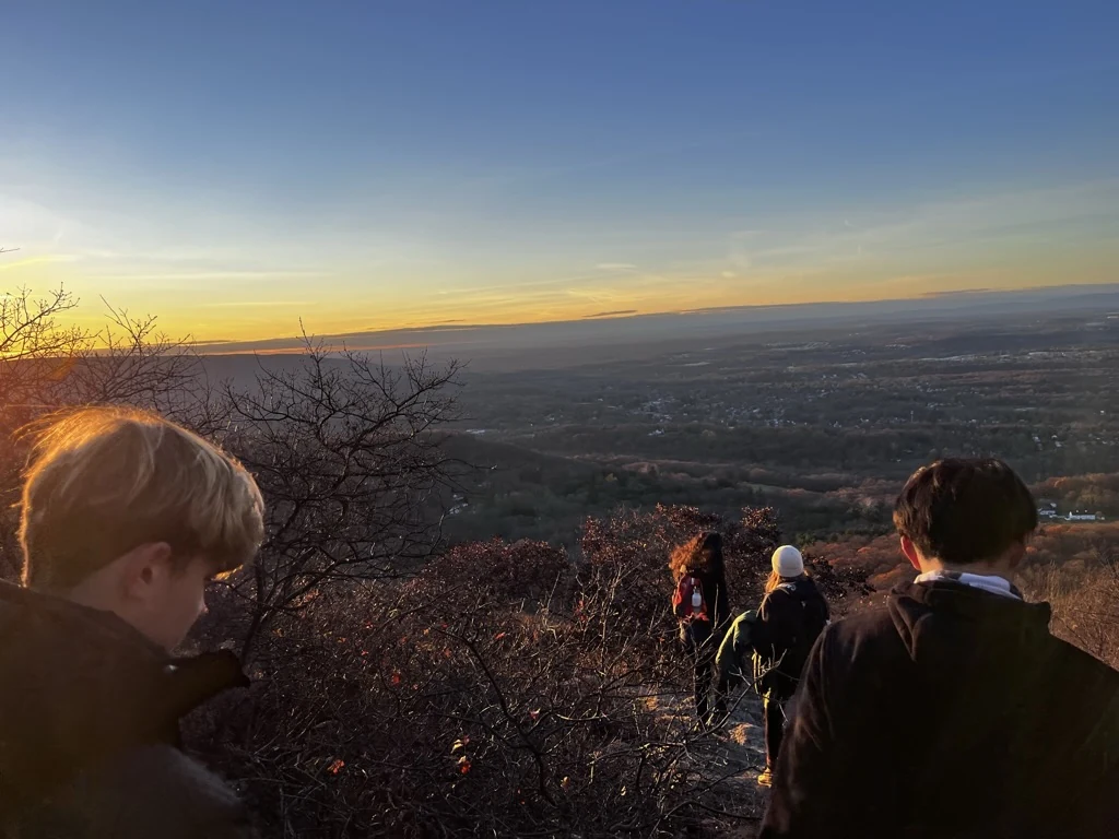 Four hikers descend a rocky trail above a valley under a blue and golden sky.