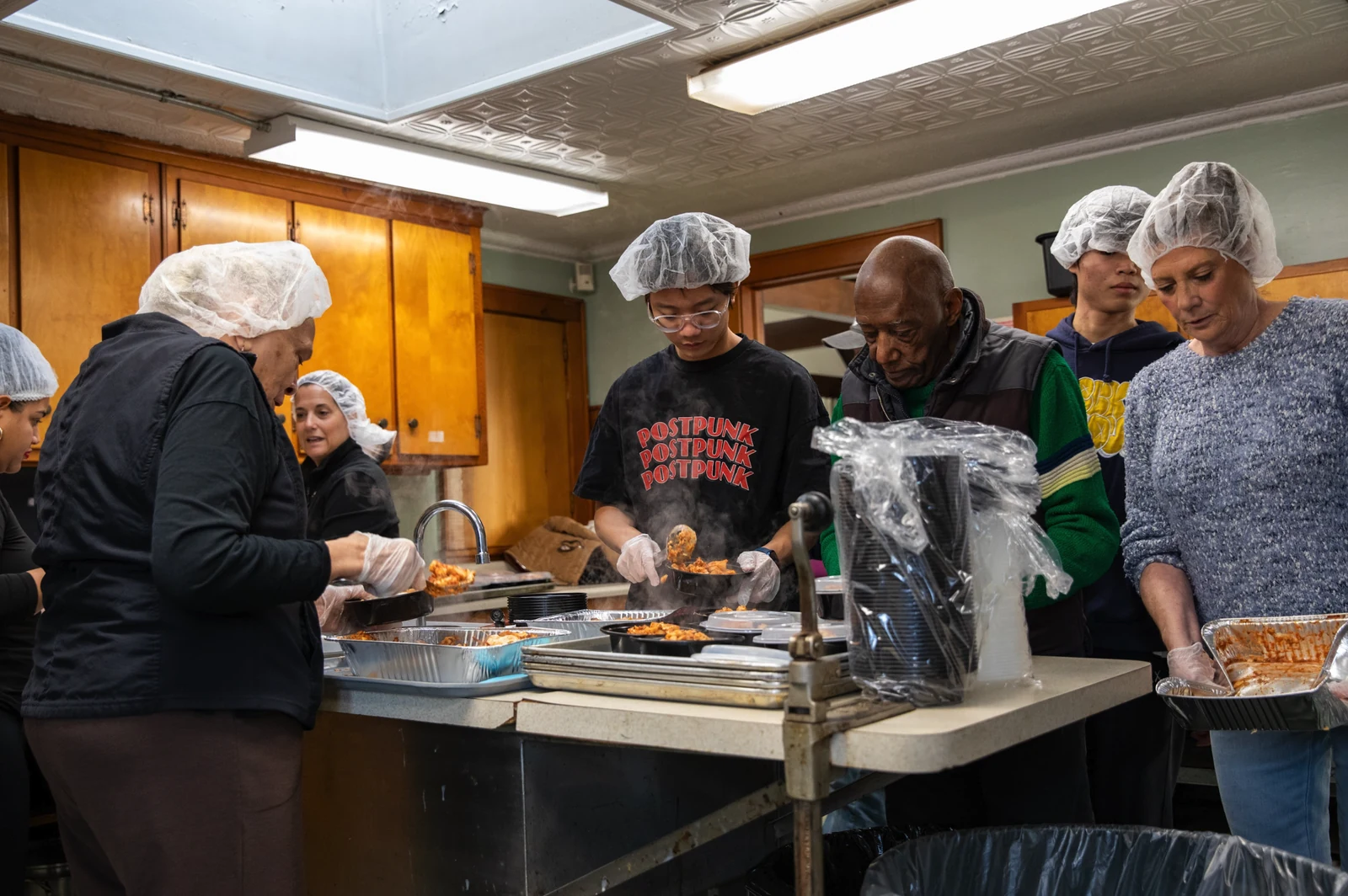 Volunteers wearing hairnets and gloves portion hot food into meal containers at a kitchen counter.