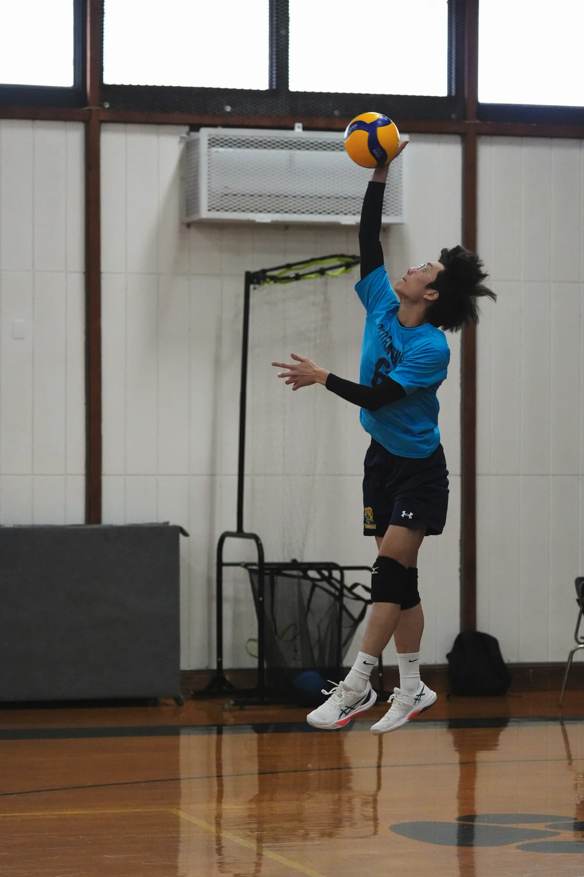A player in a blue jersey jumps toward a volleyball in an indoor gym, with the ball and full player visible.